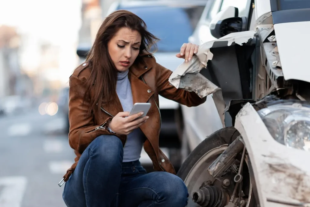 Mulher preocupada com celular na mão agachada ao lado de carro batido na rua registrando sinistro de veículo