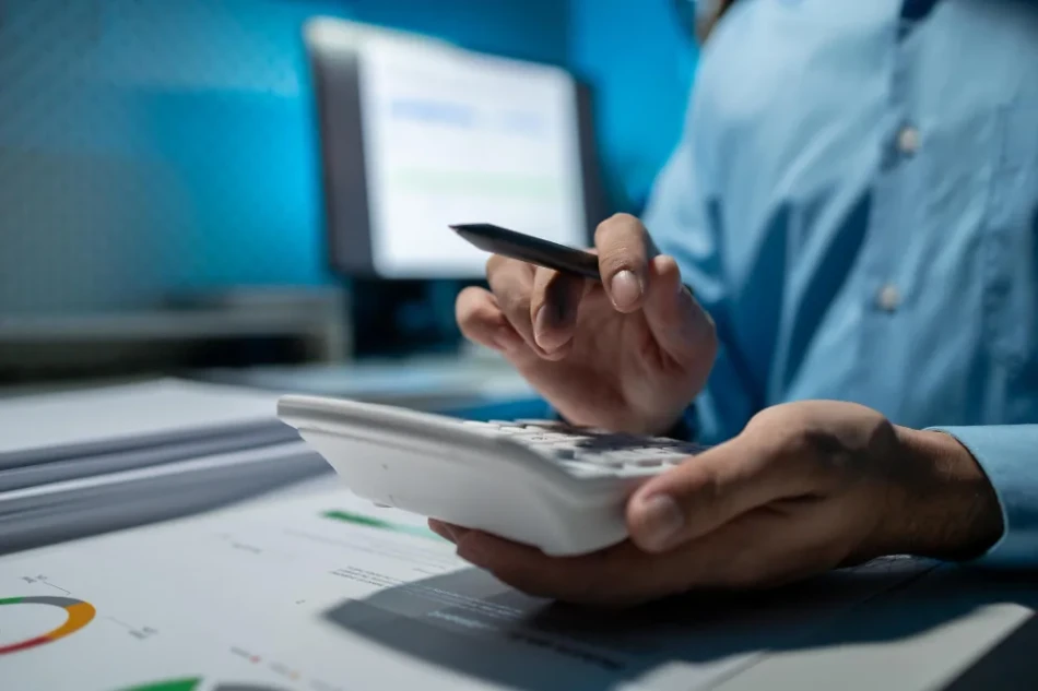Pessoa usando calculadora sobre relatorios com graficos coloridos para analise de custos operacionais com monitor ao fundo