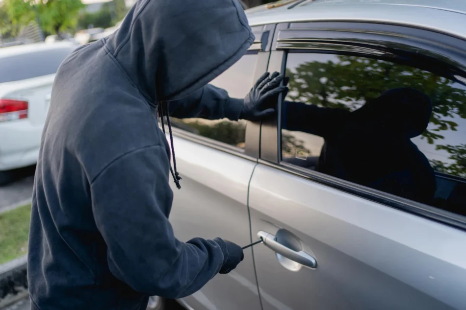 Pessoa encapuzada com luvas tentando arrombar porta de carro prata representando roubo de carros em via publica